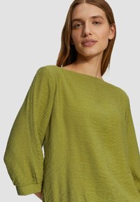 Groene, gekleurde top met een wijde hals en driekwartmouwen, gemaakt van lichtgewicht stof met een subtiele gekreukelde uitstraling.