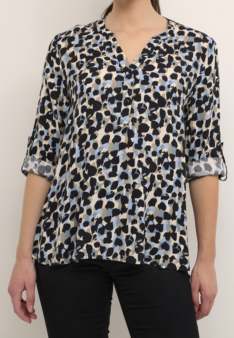 Femme portant un chemisier ample à manches longues avec un motif abstrait tacheté bleu, noir et beige, associé à un pantalon noir.