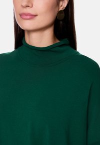 Grüner Pulli mit hohem Kragen aus weichem Stoff, mit lockerer Passform und glatter Textur. Kombiniert mit runden Ohrringen in goldfarbener Ausführung.