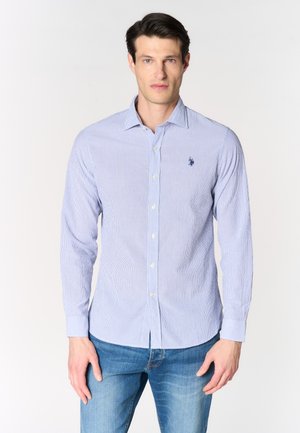 Uomo che indossa una camicia azzurro chiaro a maniche lunghe con sottili righe e logo blu ricamato sul petto, abbinata a jeans blu.