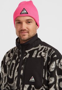 Un bonnet en tricot rose avec un logo triangulaire, associé à une veste polaire noire et grise présentant un motif texturé et une poche zippée.