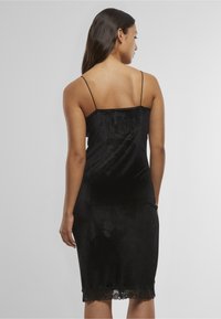 Naine, kellel on pikad tumedad juuksed, seljas must sametist lipsukleit, millel on õhukesed rihmad ja pitsist ääris, vaadatuna tagant ühetaustalise tausta ees.