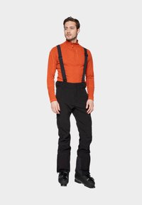 Schwarze Skihosen mit verstellbaren schwarzen Trägern, kombiniert mit einem enganliegenden orangen Langarmoberteil. Glänzende schwarze Skischuhe runden das Outfit ab.