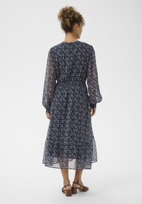 O femeie cu părul creț prins într-un coc poartă o rochie midi cu mânecă lungă, de culoare bleumarin cu flori, și sandale cu toc pătrat de culoare maronie, stând în fața unui fundal alb simplu.