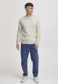 Suéter color crema con puños y dobladillo acanalados, combinado con jeans azules sueltos y zapatillas blancas, de pie contra un fondo neutro.