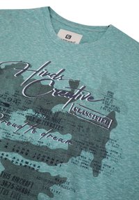 Turfärgad bomulls-T-shirt med ett texturerat grafiskt tryck som innehåller text, abstrakta former och kontrasterande färger. Rund halsringning och korta ärmar.