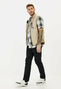 Beige gesteppte Weste über einem blauen karierten Kurzarmhemd, kombiniert mit dunklen Hosen und weißen Sneakern. Verfügt über eine Reißverschlusstasche auf der Brust.