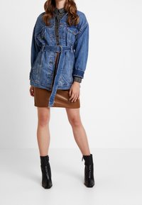 Giacca di denim con vita regolabile, dotata di due tasche frontali e cuciture a vista, indossata sopra una camicia a quadri e una gonna di pelle marrone. Stivaletti neri.