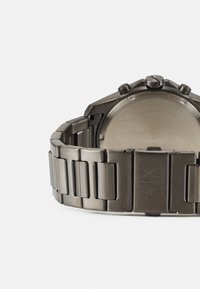 Kovové náramkové hodinky s elegantním stříbřitým páskem a úhlovým designem pouzdra. Mají hladký, neoznačený zadní kryt a několik tlačítek na vrchu.