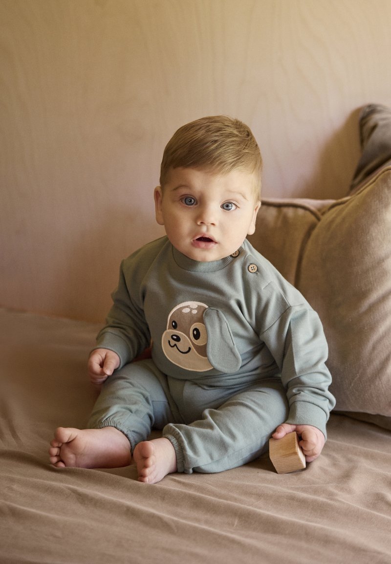 Pijama de algodón verde con un gráfico divertido de perro en la parte delantera, con cuello redondo y detalle de botones en el hombro. Textura suave.