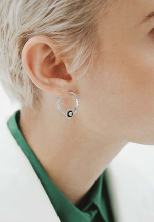 Pendiente de aro de plata con superficie texturizada, que presenta una piedra negra central y acentos claros circundantes, llevado por un modelo con cabello corto.