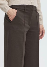 Pantalons taillés marron avec une texture lisse, dotés de poches latérales et de détails de couture subtils. Portés avec un pull en tricot de couleur claire.