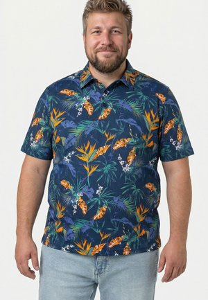 Uomo con capelli corti e barba che indossa una camicia blu a maniche corte con motivi di foglie e fiori tropicali arancioni e verdi, abbinata a jeans di denim chiaro.