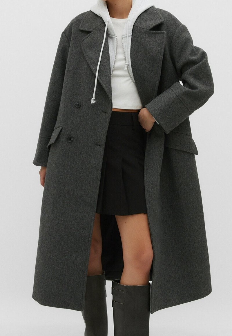 Personne portant un long manteau gris foncé croisé sur un sweat à capuche blanc, une jupe noire et des bottes hautes sombres, sur un fond uni.