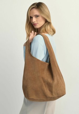 Blondynka w jasnoniebieskim swetrze niesie dużą brązową zamszową torbę na ramię, stoi lekko bokiem, patrząc w kamerę.