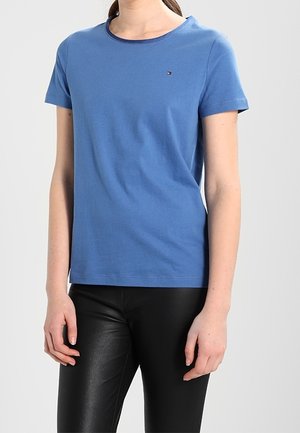 Persoon draagt een effen blauw T-shirt met korte mouwen en zwarte leerlook broek, staat met armen ontspannen langs het lichaam.
