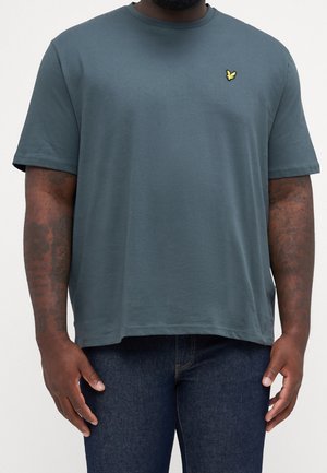 Mann trägt ein lockeres blau-grünes kurzärmeliges T-Shirt mit einem kleinen gelben Vogel-Logo auf der Brust und dunkelblaue Jeans.