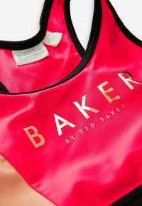 Punainen urheilullinen tanktoppi mustalla vinokaitaleella, jossa on kiiltävä kultainen "BAKER" -logo ja "by TED BAKER" -teksti. Sujuva, sileä kangas.