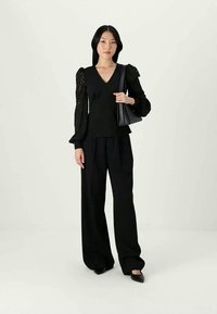 Woman wearing black sheer long-sleeve blouse, wide-leg black trousers, black pointed shoes, holding black handbag over shoulder.