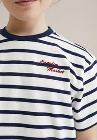 Dryžuotas marškinėliai baltais ir tamsiai mėlynais raštais, su trumpomis rankovėmis ir tamsiai mėlynu apykakle. Išsiuvinėtas užrašas "Camden Market" raudona spalva krūtinėje.