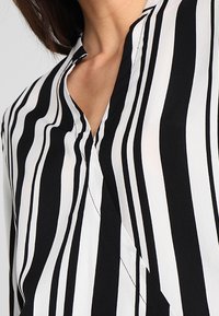 Blouse à rayures noires et blanches avec un col en V, un tissu léger et des rayures verticales marquées, présentant une texture lisse et raffinée.