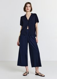 Chemise boutonnée à manches courtes bleu marine associée à un pantalon à jambes larges. Tissu lisse, coupe décontractée, et sandales plates noires avec des accents dorés.