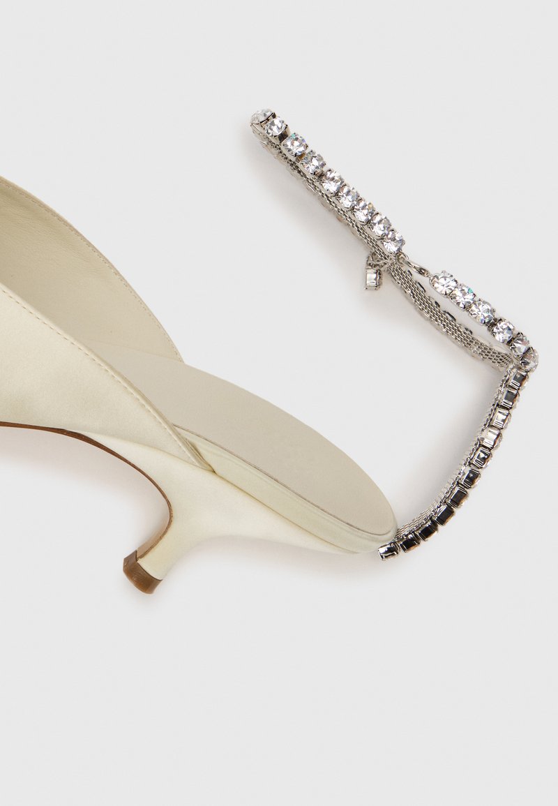 Cremefarvet satin stiletto-sko med kitten hæl, parret med et sølvfarvet rhinestone armbånd på en hvid baggrund.