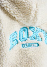 Tela de vellón esponjosa color crema con bordado "ROXY" en azul claro y acento "EST. 1990". Textura suave y bordes redondeados.
