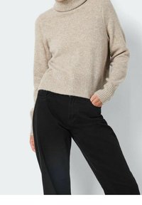 Pull à col roulé côtelé beige associé à un pantalon large taille haute noir, une main dans la poche, sur un fond uni.