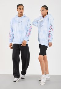 Duas modelos a usar capuzes azul claro com mangas floridas em rosa; uma com calças largas pretas e ténis, a outra com calções pretos e sapatilhas brancas de cano alto.