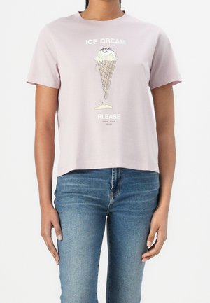 Kobieta ubrana w jasnoróżowy t-shirt z grafiką topniejącego rożka lodowego i napisem „ICE CREAM PLEASE” w połączeniu z niebieskimi dżinsami.