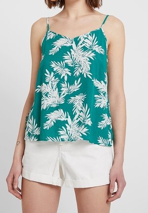 Femme portant un haut sans manches turquoise avec des motifs de feuilles blanches et un short blanc à ourlet roulé, debout devant un fond uni.