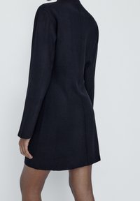 Robe en tricot noire avec une silhouette ajustée, des manches longues, un ourlet côtelé et un col montant, présentant un design minimaliste et un dos sans coutures.
