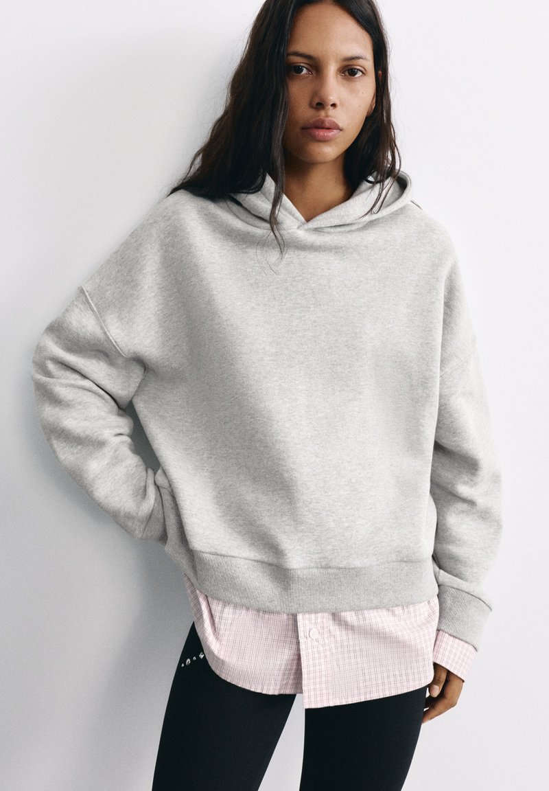 Femme aux longs cheveux foncés portant un sweat à capuche ample gris clair sur une chemise à carreaux rose et un pantalon noir, debout contre un mur blanc.