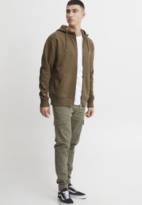 Brun zip-up hettejakke med snorer, brukt over en hvit skjorte, kombinert med olivenfargede cargo-bukser og svarte joggesko.