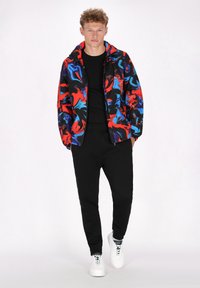 Kleurvolle, abstracte gestreepte zip-up jack in rood, blauw en zwart gedragen over een zwart shirt en zwarte joggers; gecombineerd met witte sneakers.