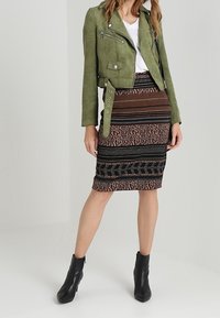 Olka vihreä mokkana oleva cropped-takki, jossa on hopeiset yksityiskohdat, yhdistettynä moniväriseen kuviolliseen kynähameeseen ja mustiin nilkkureihin.