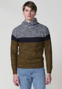 Gebreide trui met een col; heeft navy, grijs en mosterdstrepen. Textiel met structuur en een geribbelde zoom. Draagt met lichtblauwe jeans.