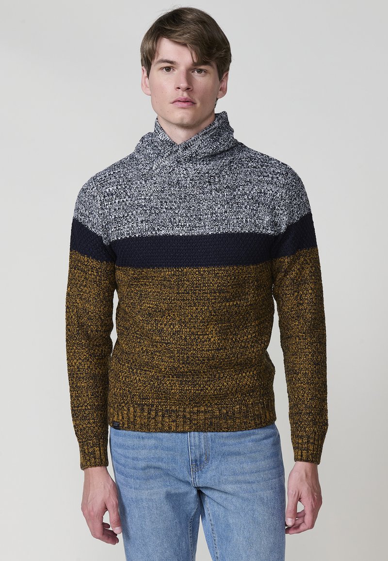 Gebreide trui met een col; heeft navy, grijs en mosterdstrepen. Textiel met structuur en een geribbelde zoom. Draagt met lichtblauwe jeans.