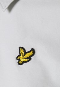 Biała teksturowana tkanina z haftowanym logo orła w kolorze czarnym i żółtym po lewej stronie. Prosty design koszulki polo.