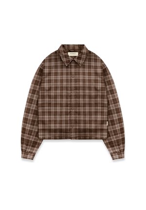 Mann trägt eine braune karierte Knopfleistenjacke und ausgewaschene graue Jeans und steht vor einem schlichten hellen Hintergrund.