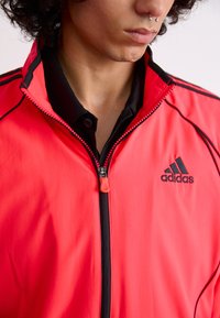 Red lightweight zip-up jacket with black accents and adidas logo. Features a high collar and smooth texture. Zipper pull with accent color.