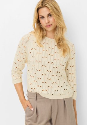 Blond kvinde iført en cremefarvet strikket sweater med ovale udskæringer og beige højtaljede bukser poserer med den ene hånd i lommen.