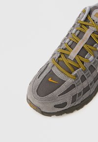 Pilka sportinė avalynė su tekstūruotu zomšiniu ir tinklinio viršumi, ryškiai geltonos spalvos raišteliai, išraiškingos siūlės ir mažas oranžinis logotipas ant pirštų dalies.