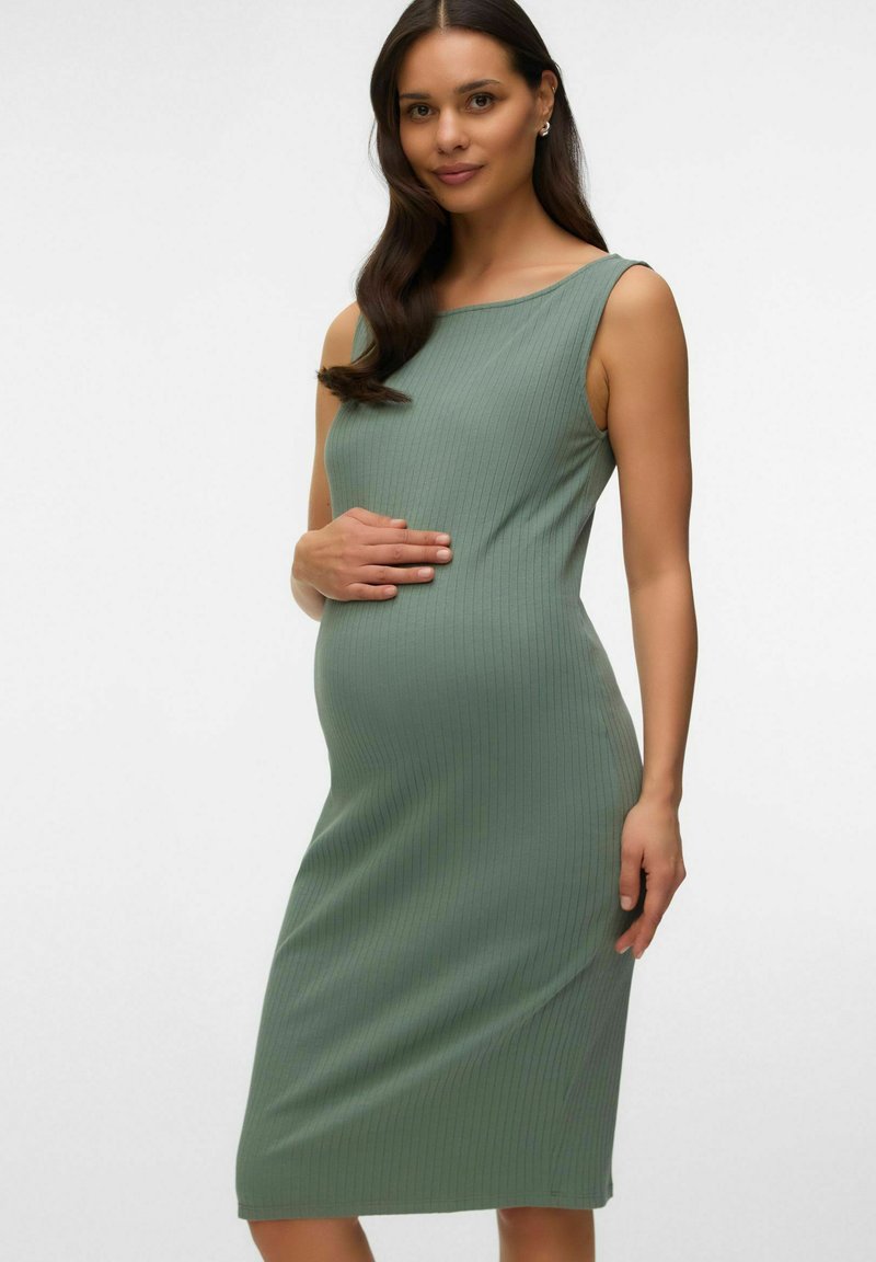 Femme enceinte aux longs cheveux foncés portant une robe verte côtelée sans manches, tenant doucement son ventre, debout devant un fond uni.