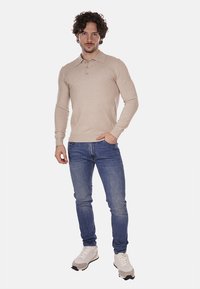 Polo beige a maniche lunghe con una chiusura a tre bottoni, abbinato a jeans blu e sneakers bianche. Design semplice e aderente con tessuto testurizzato.