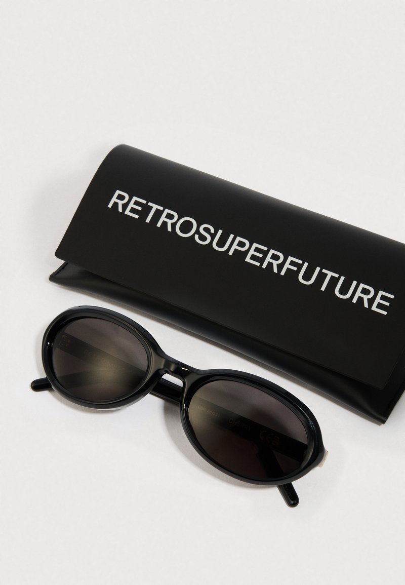Des lunettes de soleil ovales noires avec des verres foncés, présentées à côté d'une pochette noire mate arborant le texte "RETROSUPERFUTURE" en lettres blanches.