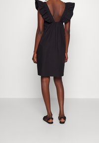 Schwarzes Kleid mit Rüschenärmeln, offener Rückseite, knielangem Saum und weichem Material. Getragen mit flachen schwarzen Sandalen.
