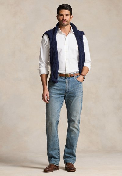 Mann steht mit der Hand in der Tasche, trägt ein weißes Hemd mit Knöpfen, blaue Jeans, braune Schuhe und einen marineblauen Pullover über den Schultern.