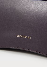 Sac en cuir violet foncé, présentant une texture lisse avec un logo "Coccinelle" en doré. Comprend des coutures détaillées le long des bords.
